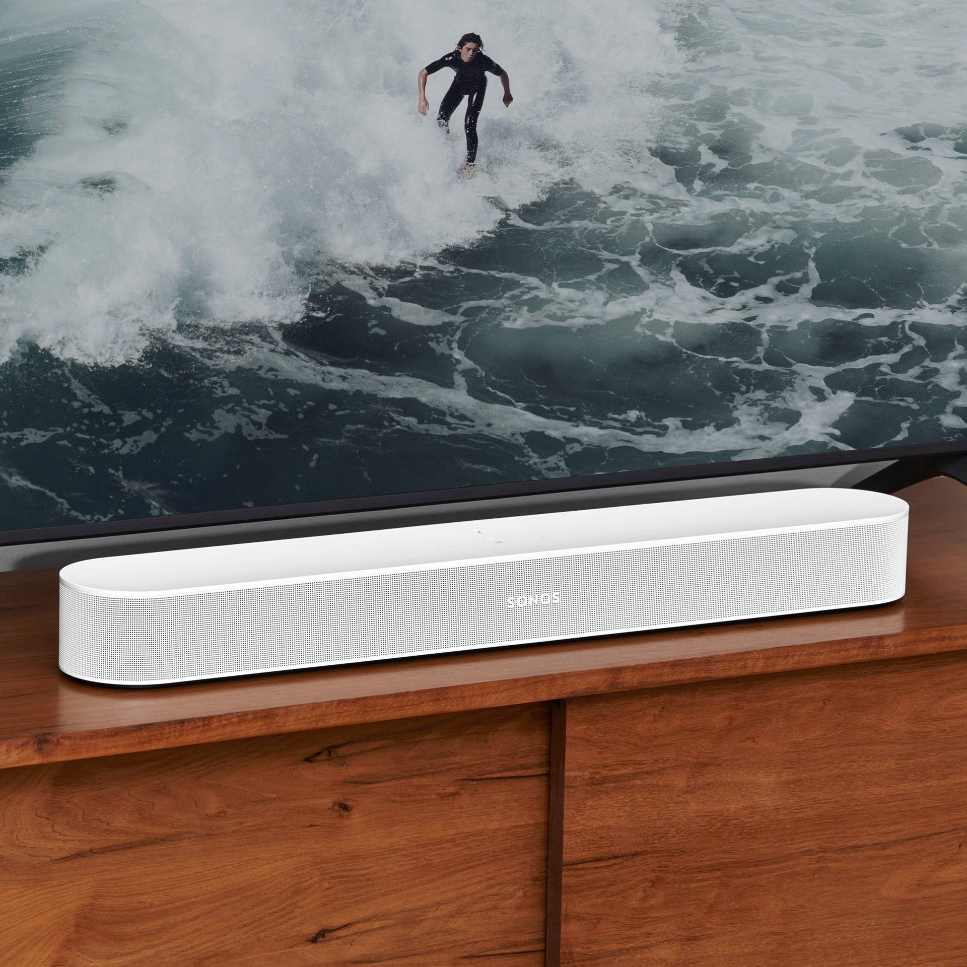 Sonos soundbar on a wooden surface with a television displaying a surfer on a wave in the background