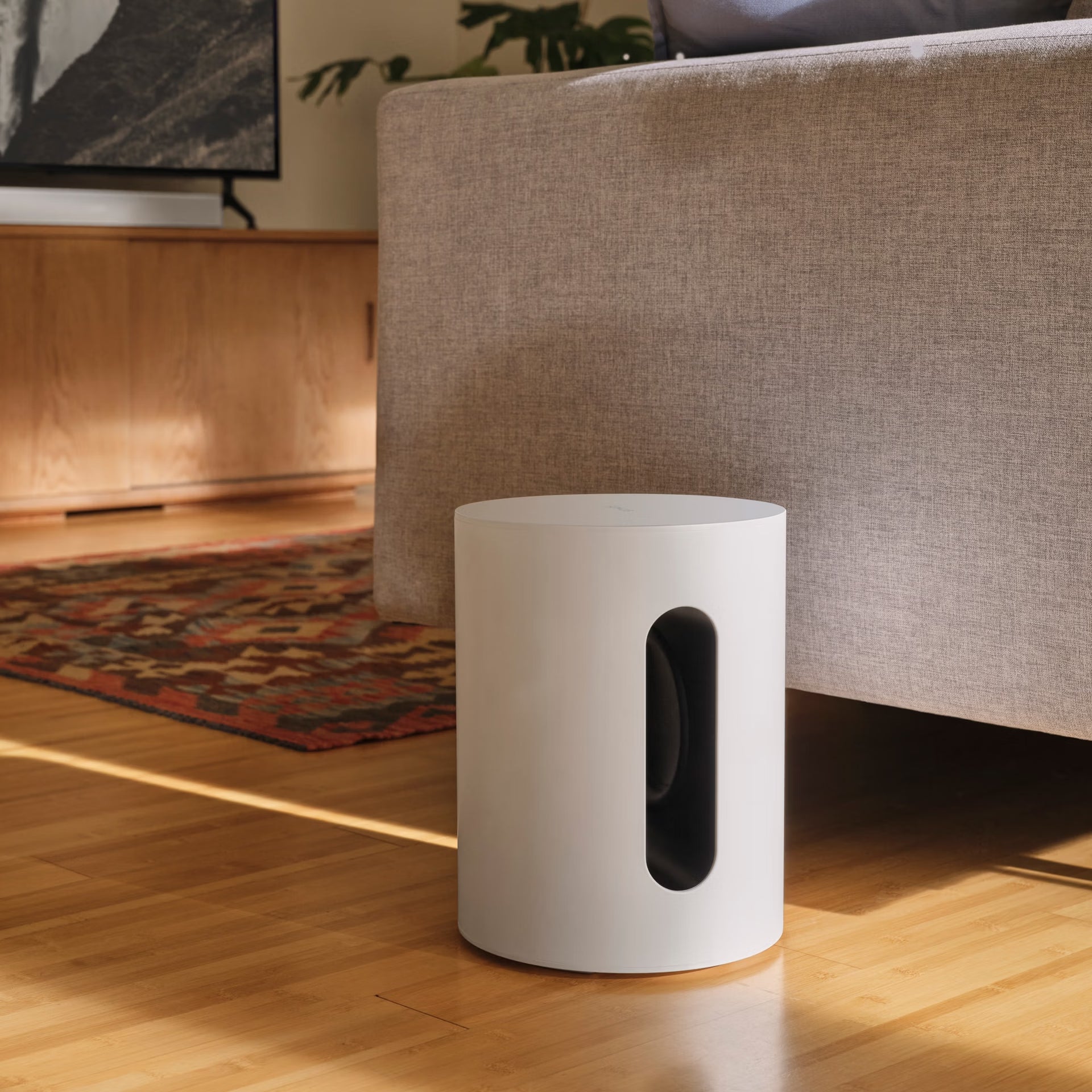 Sonos Sub Mini white compact smart subwoofer  on a wooden floor with a couch and television in the background
