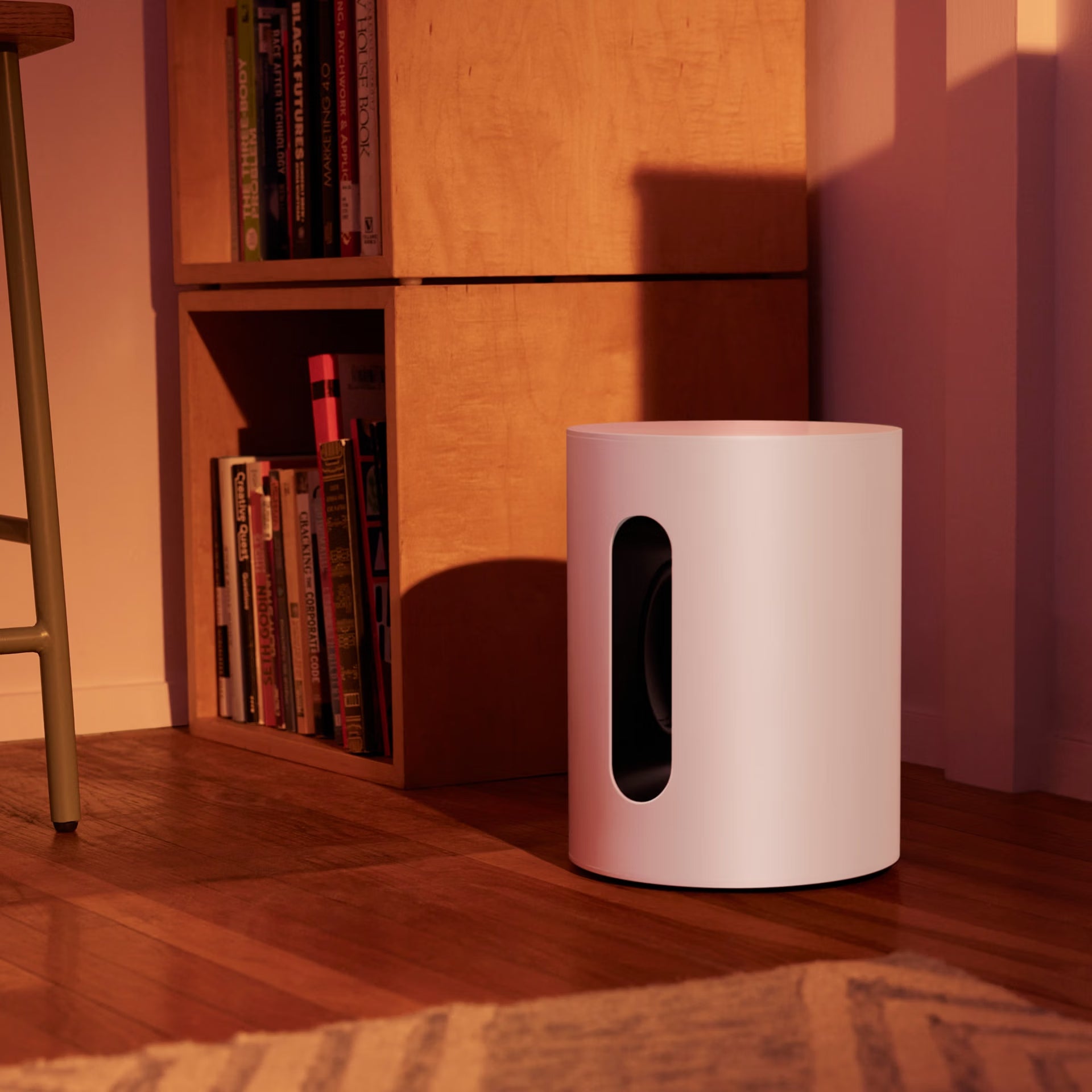 Sonos Sub Mini white compact smart subwoofer with a black circular cutout on a wooden floor next to a bookshelf.