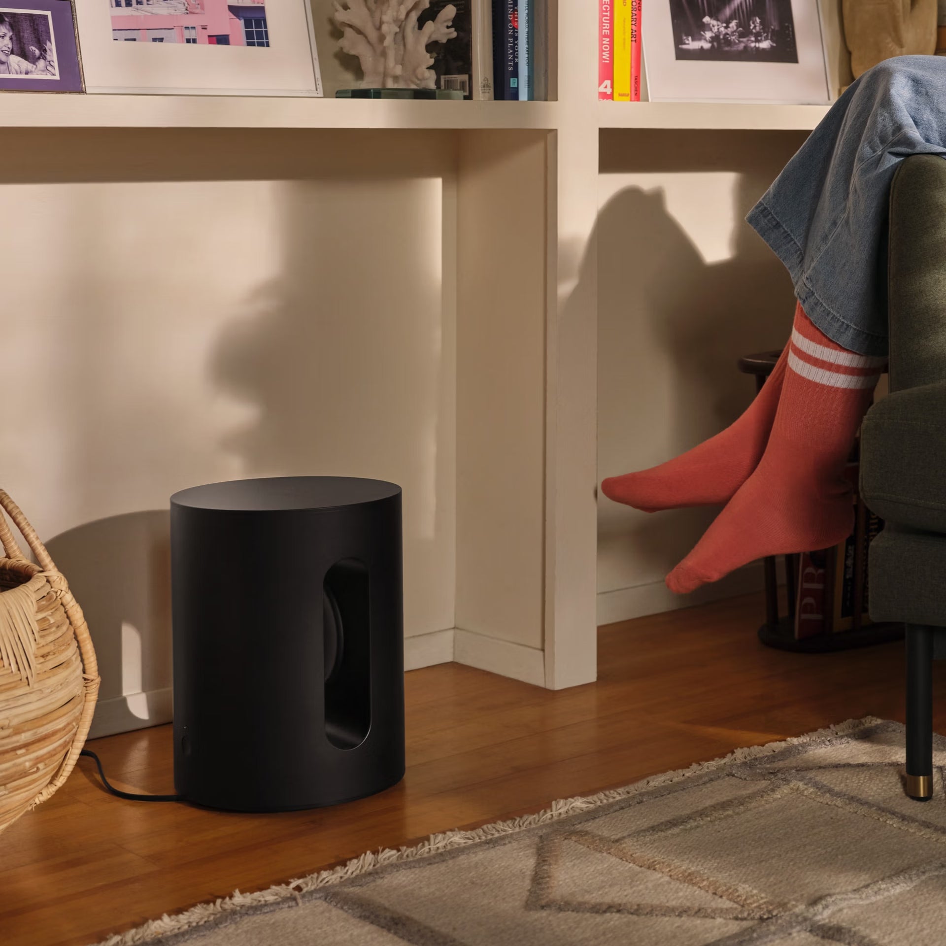 Sonos Sub Mini black subwoofer on a wooden floor with a person sitting on a chair wearing red socks.
