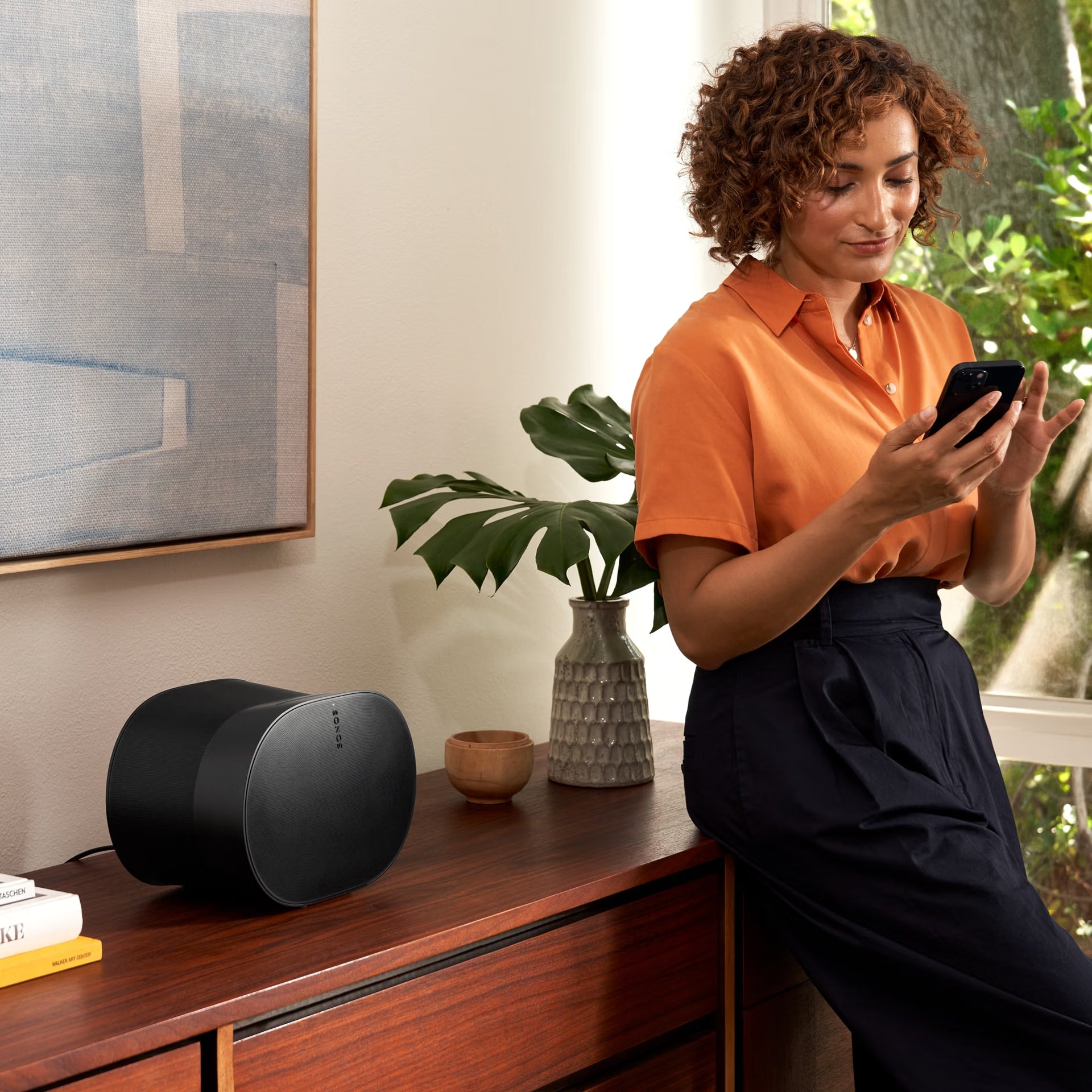 Woman using a smartphone in a living room with a Sonos Era 300 UK (Black) speaker and plant on a wooden cabinet.