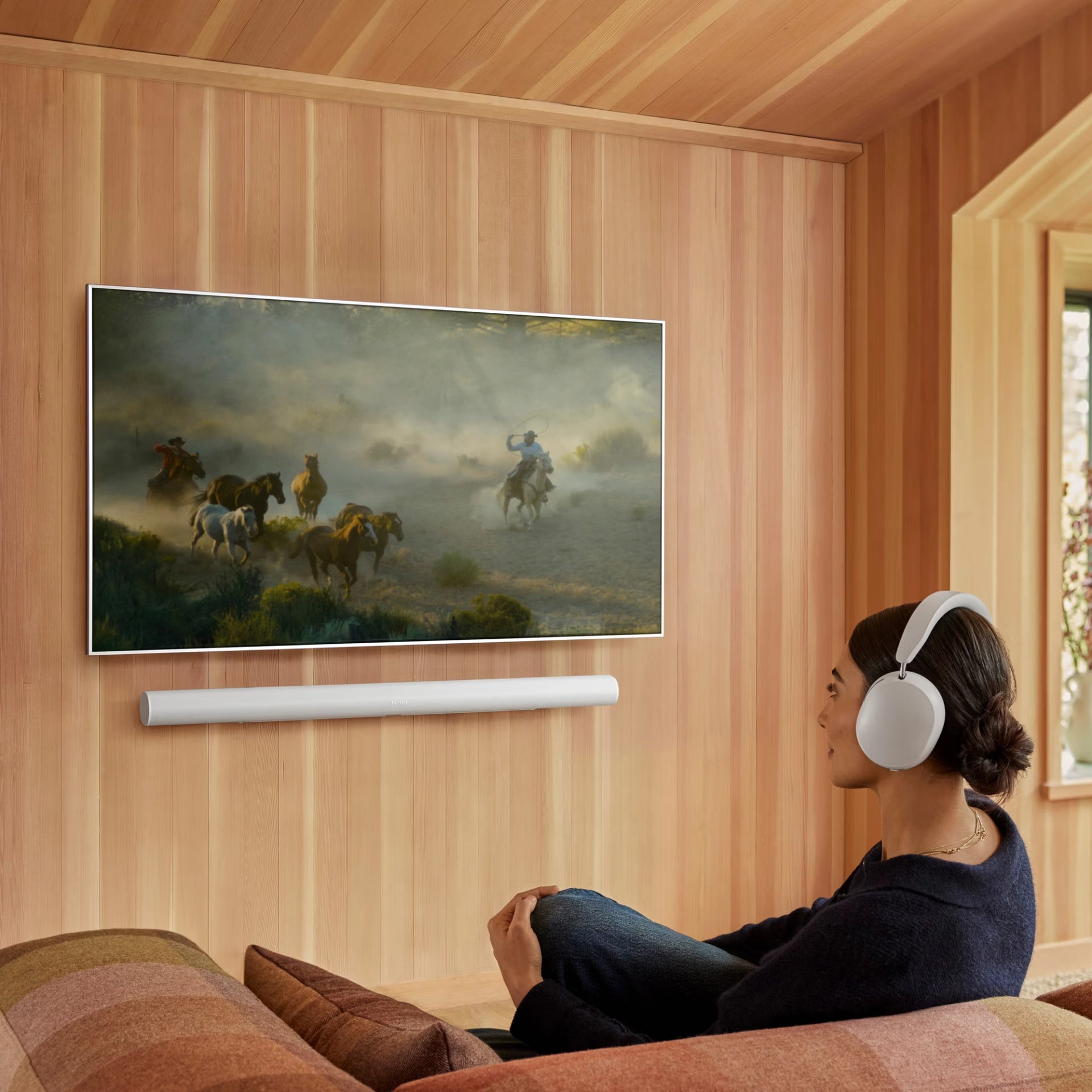 Person sitting on a couch wearing headphones, watching a painting of cowboys on a screen.

