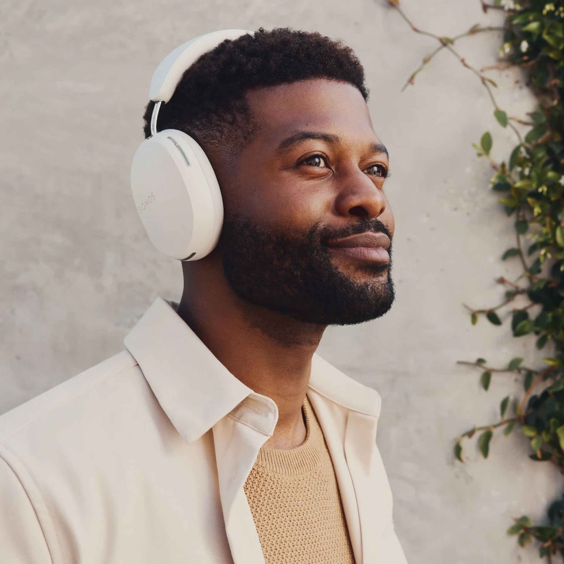 Man wearing Sonos Ace white headphones, smiling outdoors with a neutral wall and greenery in the background
