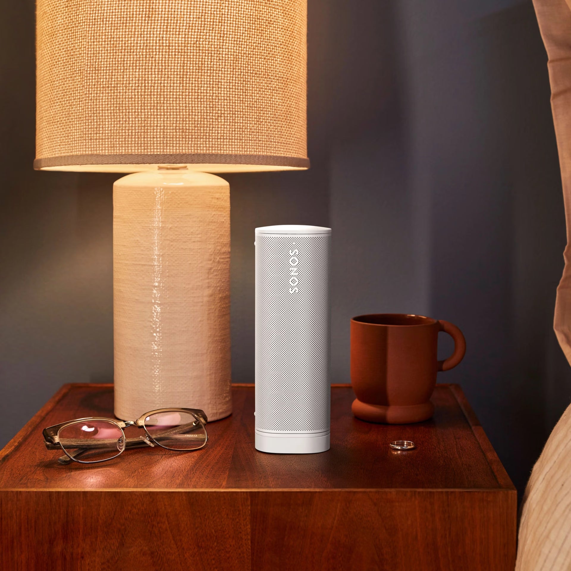 Nighstand with lamp, Sonos Roam2 WHITE speaker, mug, and glasses on a wooden surface.