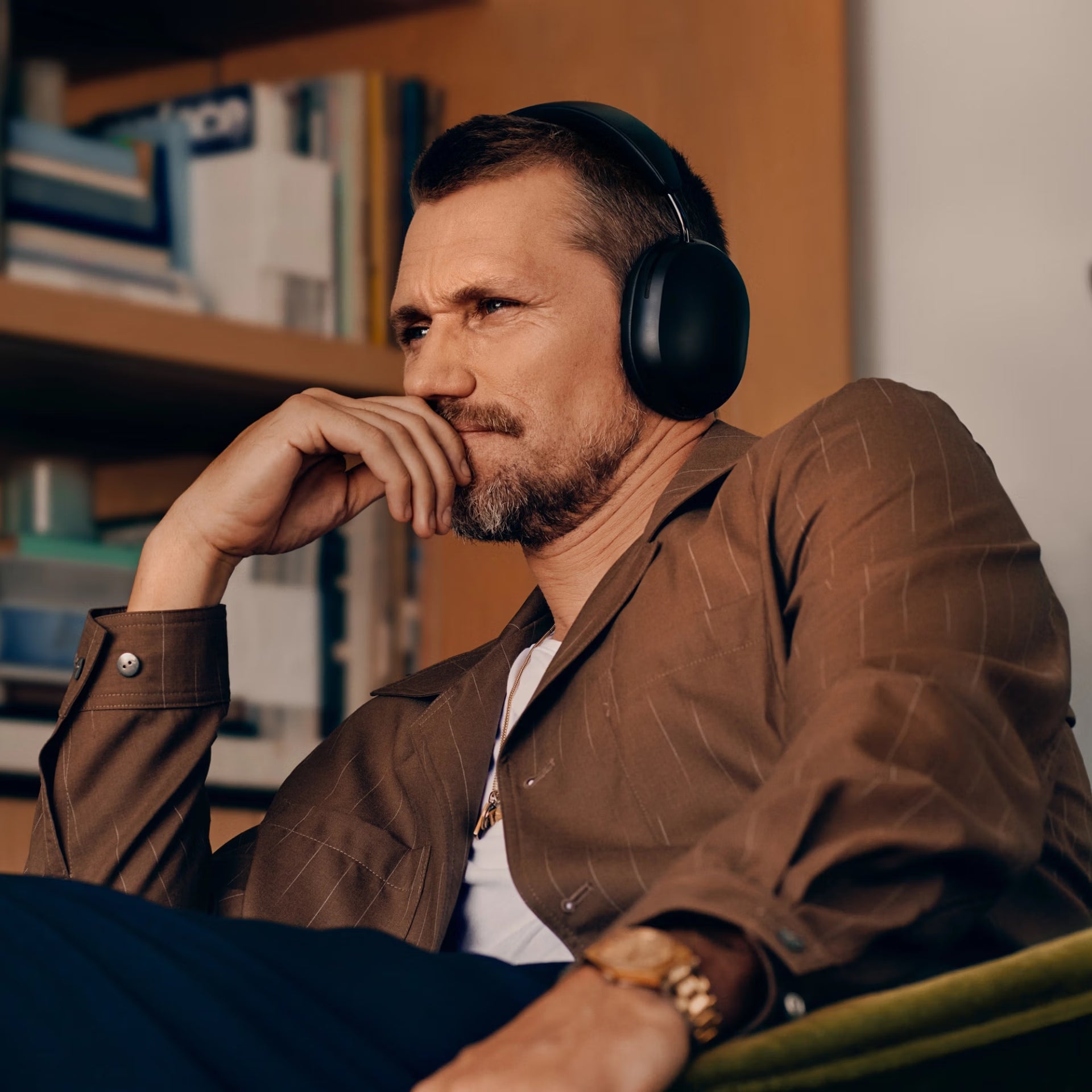 Man wearing SonosAce_Black headphones sitting on a couch in a room with bookshelves.