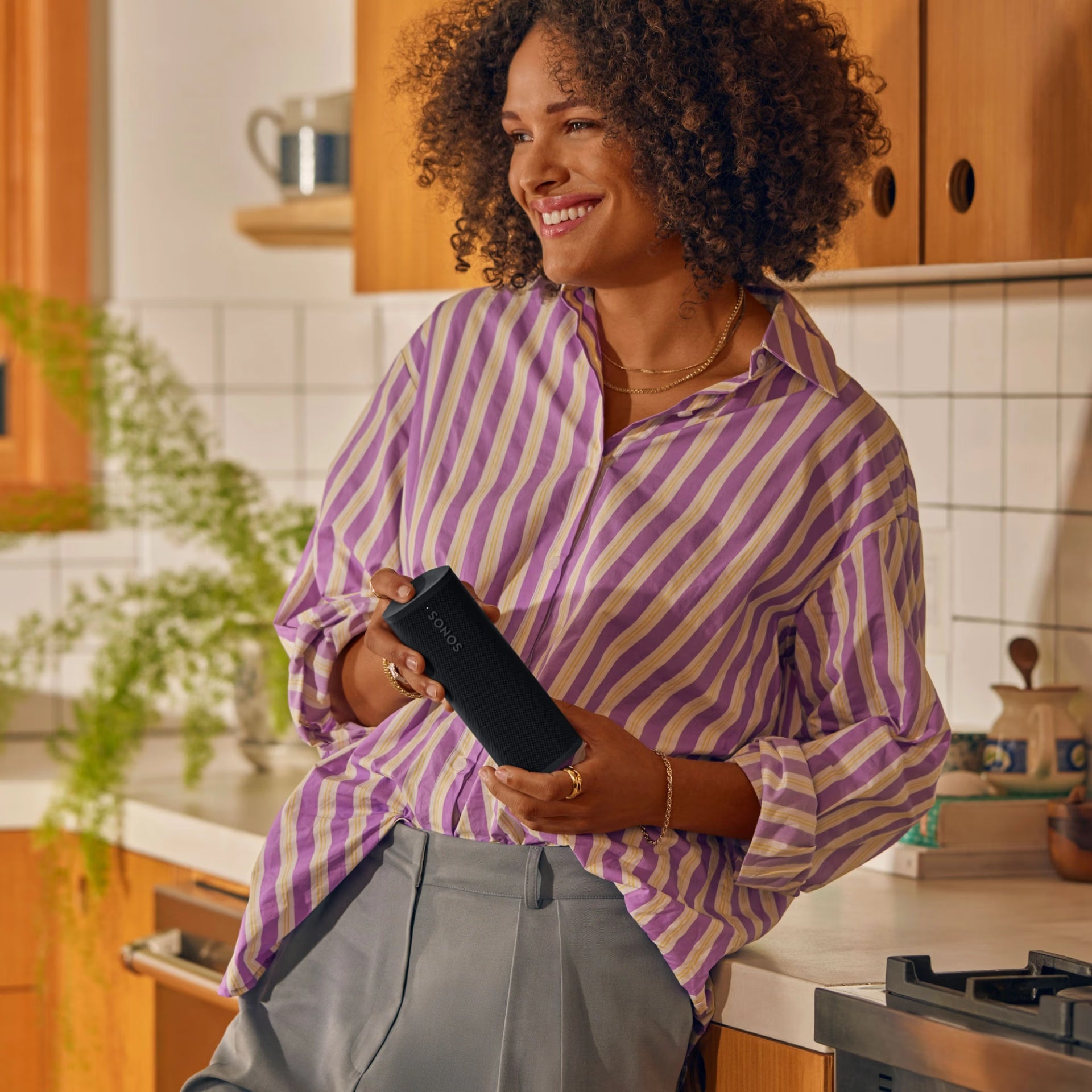 Woman in a kitchen holding a Sonos Roam 2 (Black) , wearing a purple and yellow striped shirt.